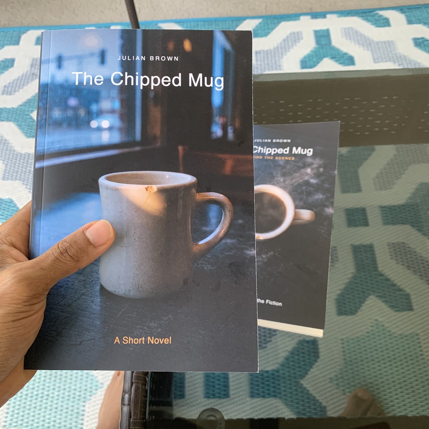 A hand holding The Chipped Mug with its companion volume nearby